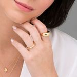 Mão de mulher com cabelo escuro usando dois anéis dourados grandes e uma pequena corrente dourada com pingente, vestindo blusa branca.