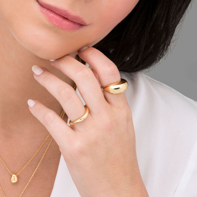 Mão de mulher com cabelo escuro usando dois anéis dourados grandes e uma pequena corrente dourada com pingente, vestindo blusa branca.