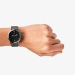 Relógio preto com pulseira de malha metálica no pulso de uma mão, exibindo mostrador escuro com detalhes em branco.