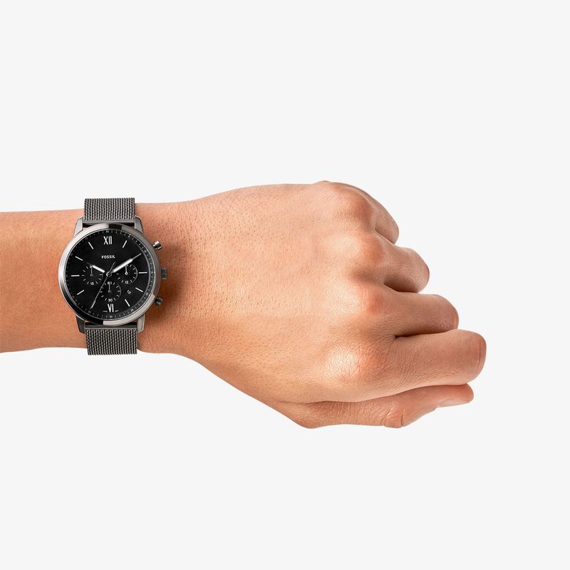 Relógio preto com pulseira de malha metálica no pulso de uma mão, exibindo mostrador escuro com detalhes em branco.