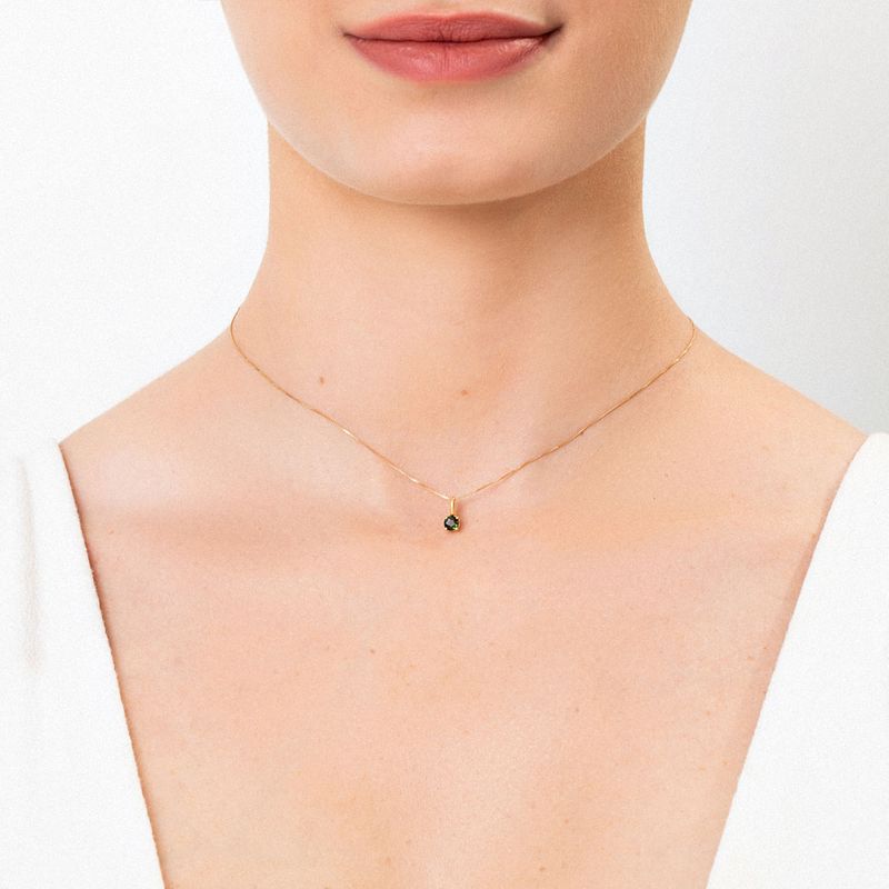 Colar delicado de corrente dourada com pingente pequeno negro, usado por uma pessoa com pele clara e vestido branco.