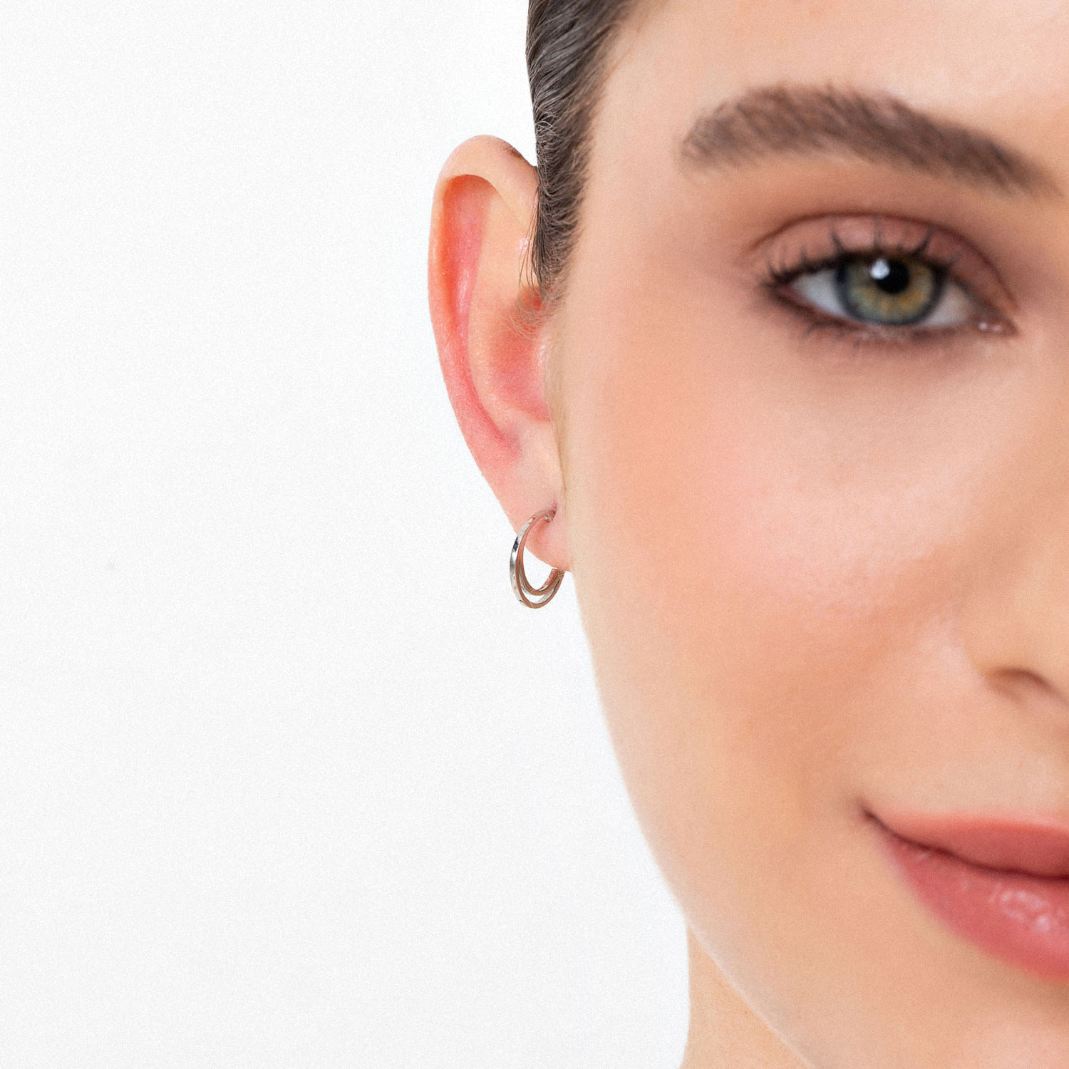 Close do rosto de uma mulher mostrando seus olhos, orelha com pequenos brincos de argola e parte da bochecha e lábios, com pele lisa e maquiagem.