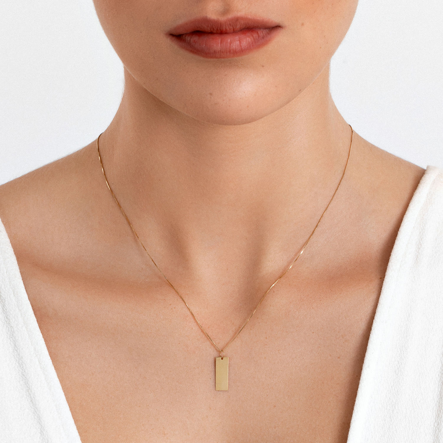Colar dourado delicado com pingente retangular, usado por uma mulher com pele clara e camisa branca.