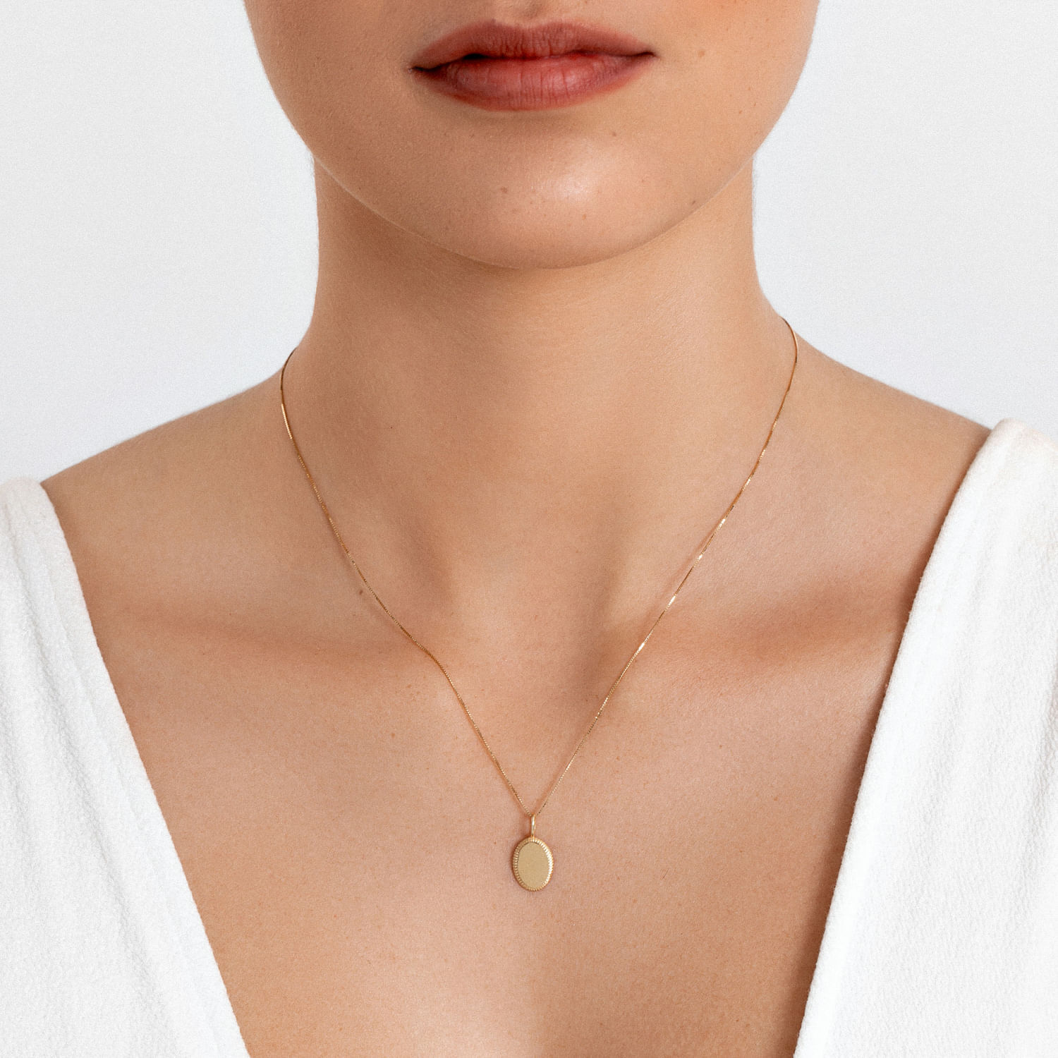 Colar dourado com pingente oval sólido, usado por uma mulher com camiseta branca e pele clara.