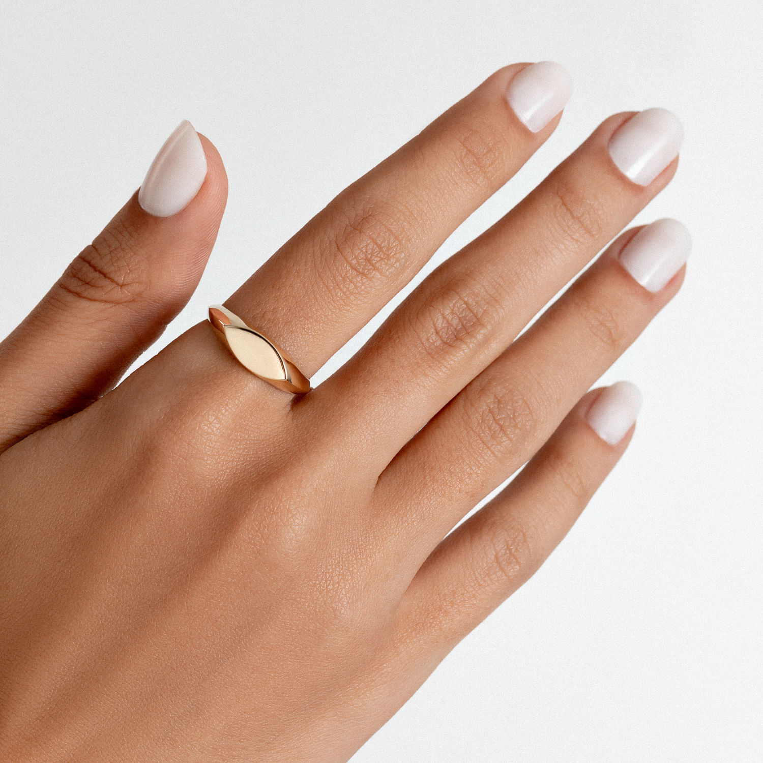 Mão com unhas brancas e um anel dourado em formato de folha na dedo médio, contra fundo branco.