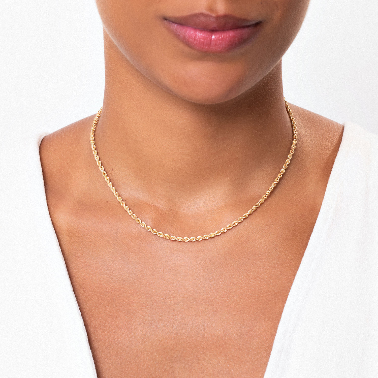 Colar de corrente dourada delicada e fina, usado no pescoço de uma mulher com pele clara, vestindo blusa branca.