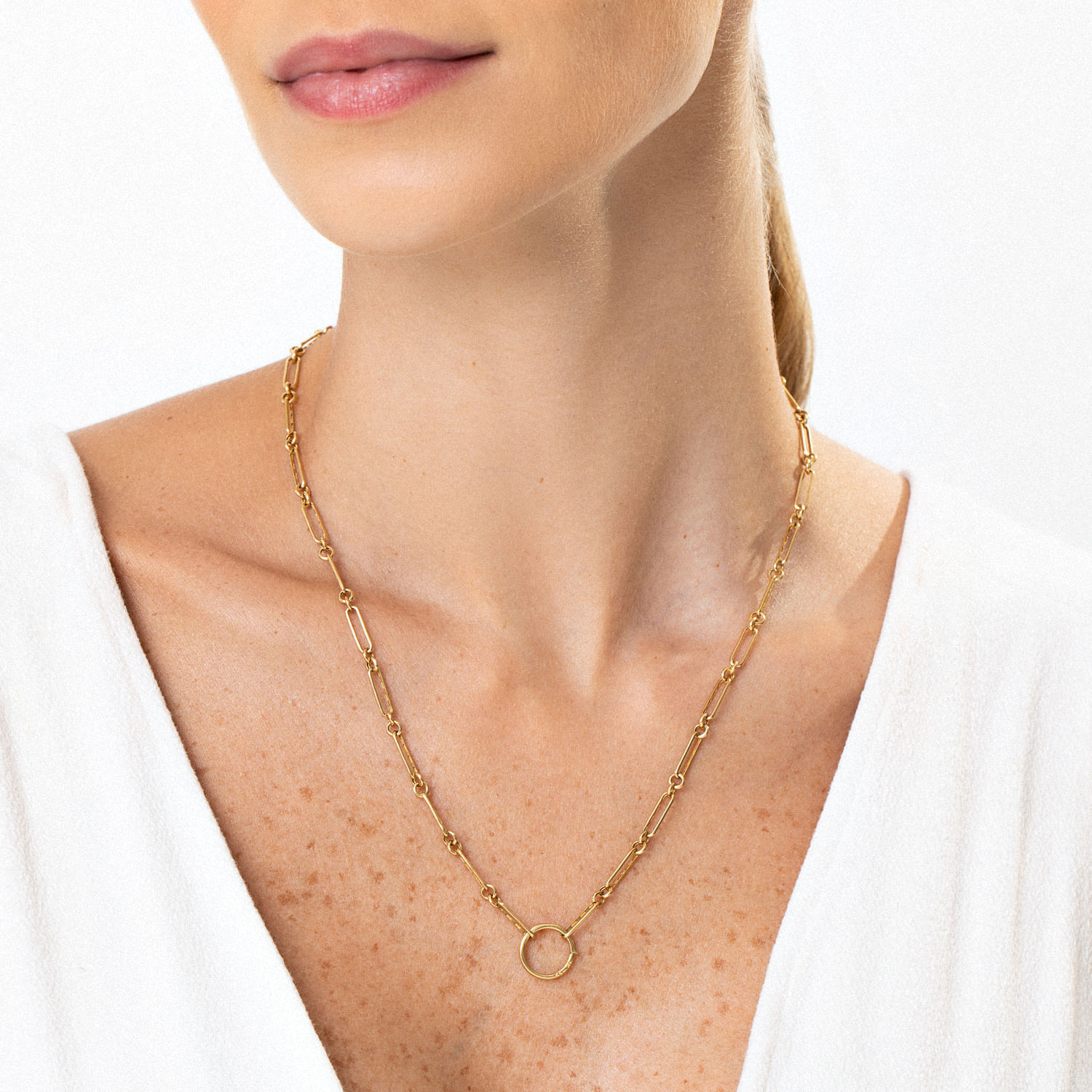 Colar dourado com elos longos e um círculo na frente, usado por uma mulher com pele clara e blusa branca.