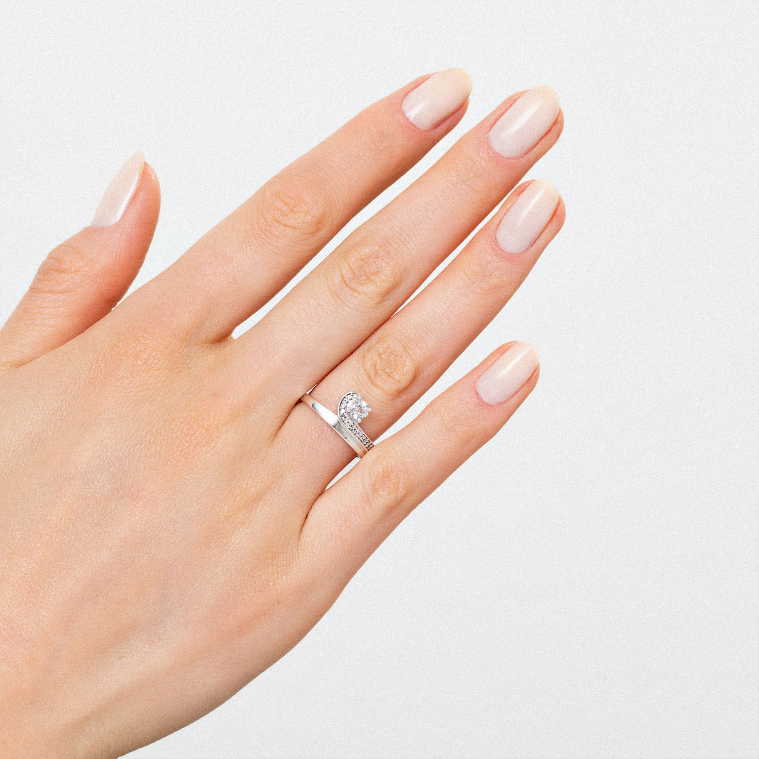 Dedos de uma mão com unha pintada de cor nude e anel prateado com diamantes, exibindo a mão contra fundo branco.