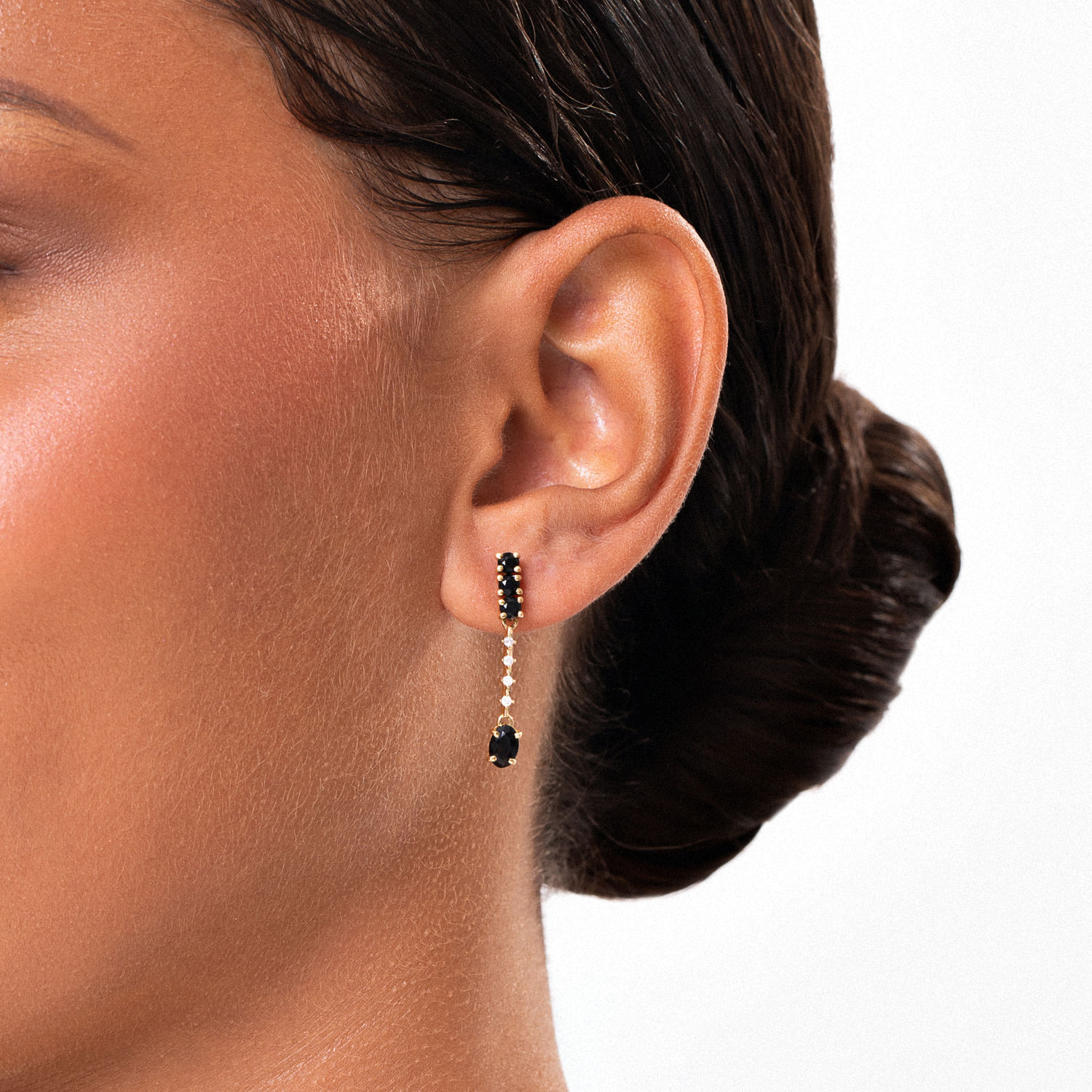 Brinco de argola longo com pedra preta e detalhes dourados na orelha de mulher com cabelo preso.