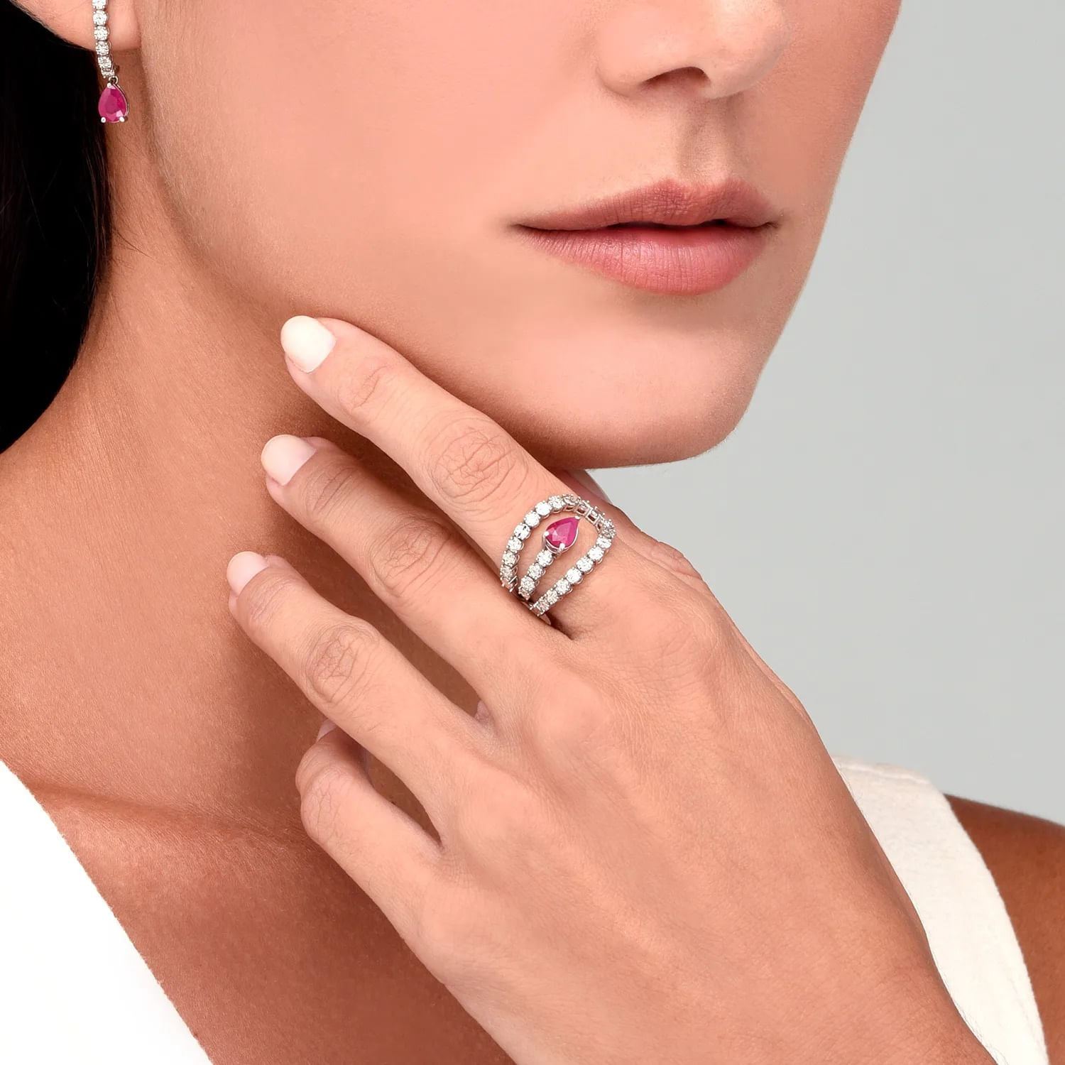 Mão de uma mulher com anel e brincos de joias com pedras rosas e diamantes, próxima ao rosto, com fundo cinza claro.
