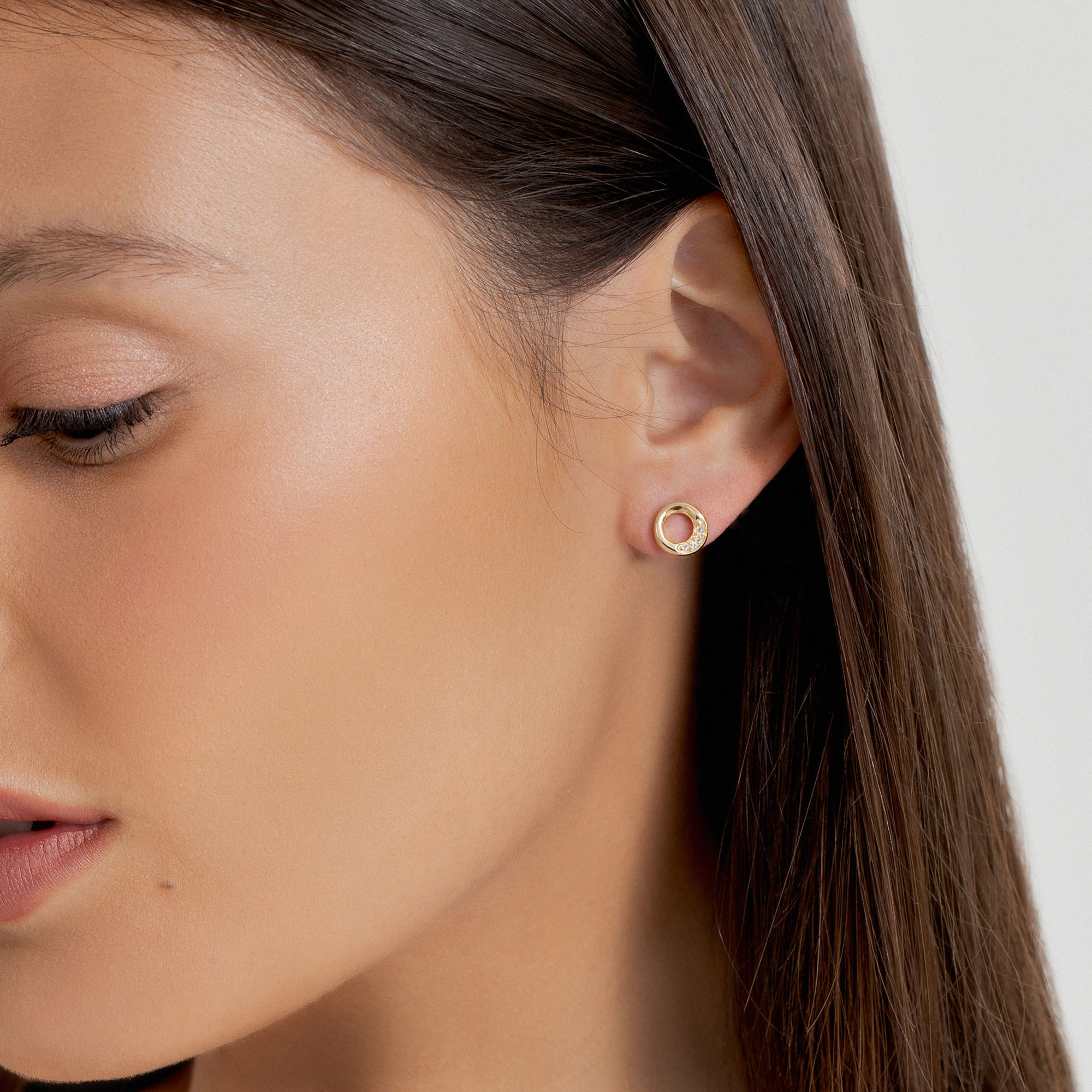 Close do rosto de uma mulher mostrando sua orelha com um brinco circular dourado e rosa, pele lisa e cabelo castanho.