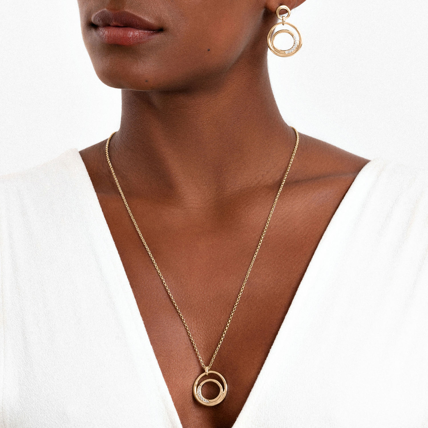 Colar e brincos de argola dourados com círculos entrelaçados, usados por uma mulher com pele escura e blusa branca.