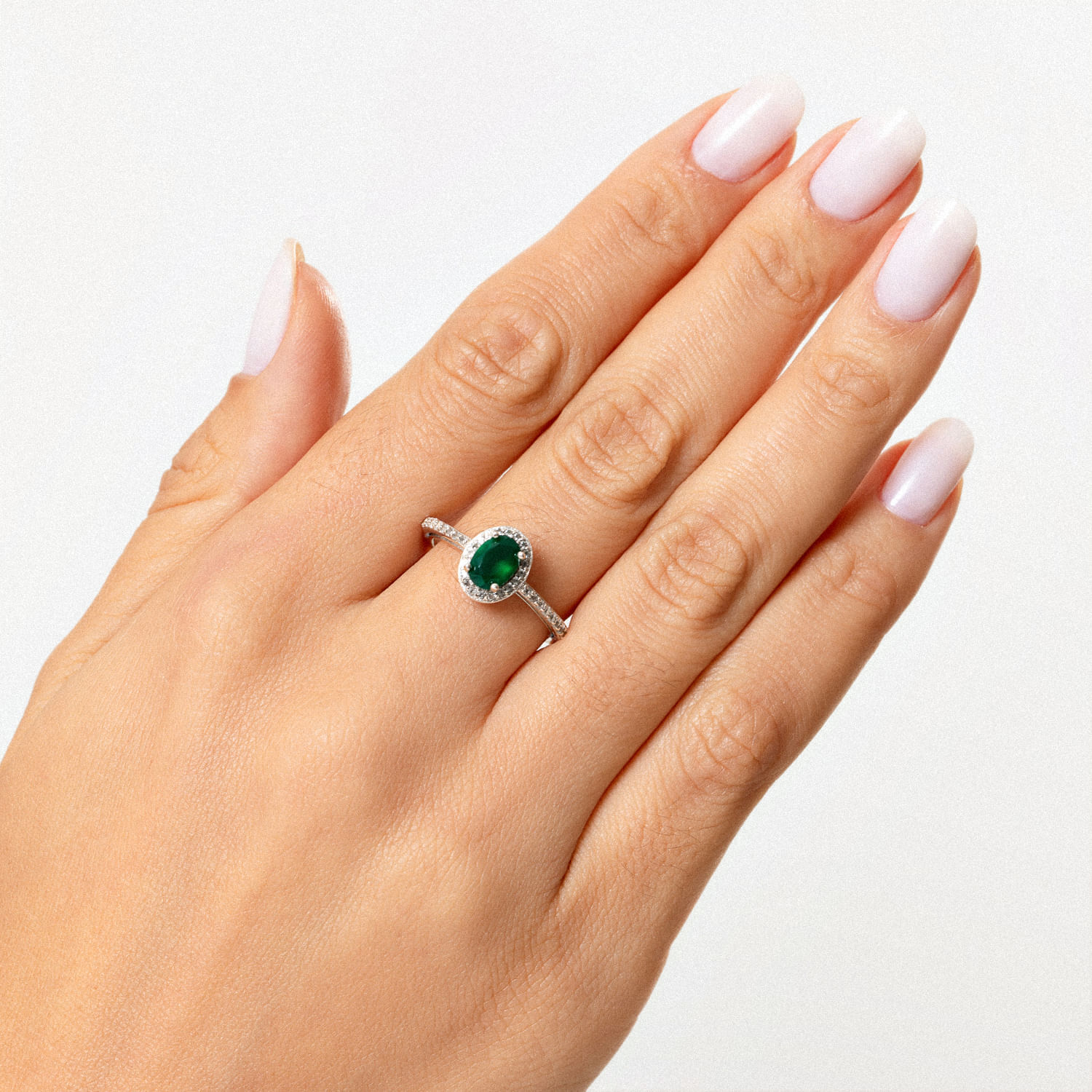 Anel com pedra oval verde e detalhes em cristais brancos em um dedo de mão feminina com unhas pintadas de branco.