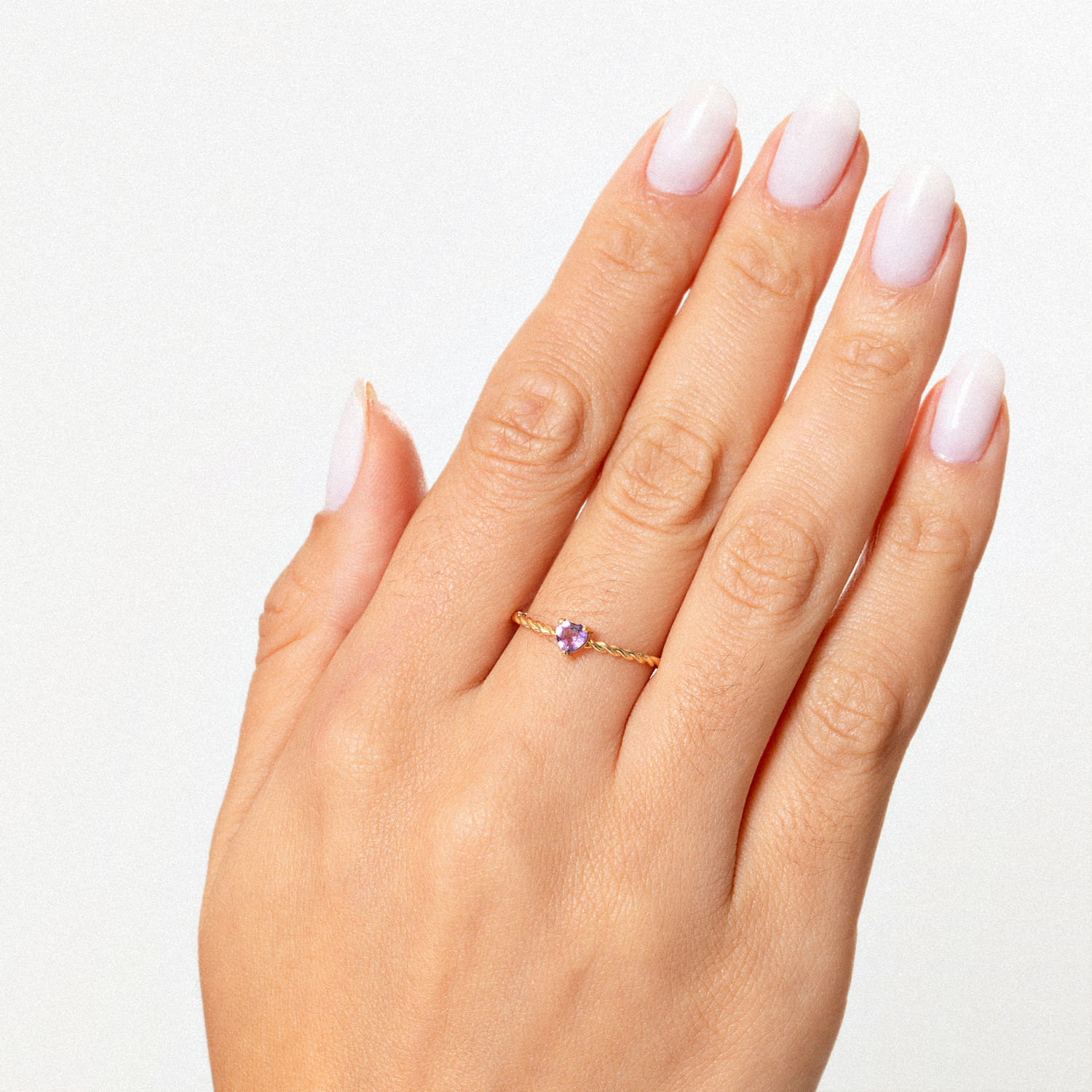 Dedo com unha esmaltada de branco e usando um anel delicado com pedra roxa na mão esquerda sobre fundo branco.