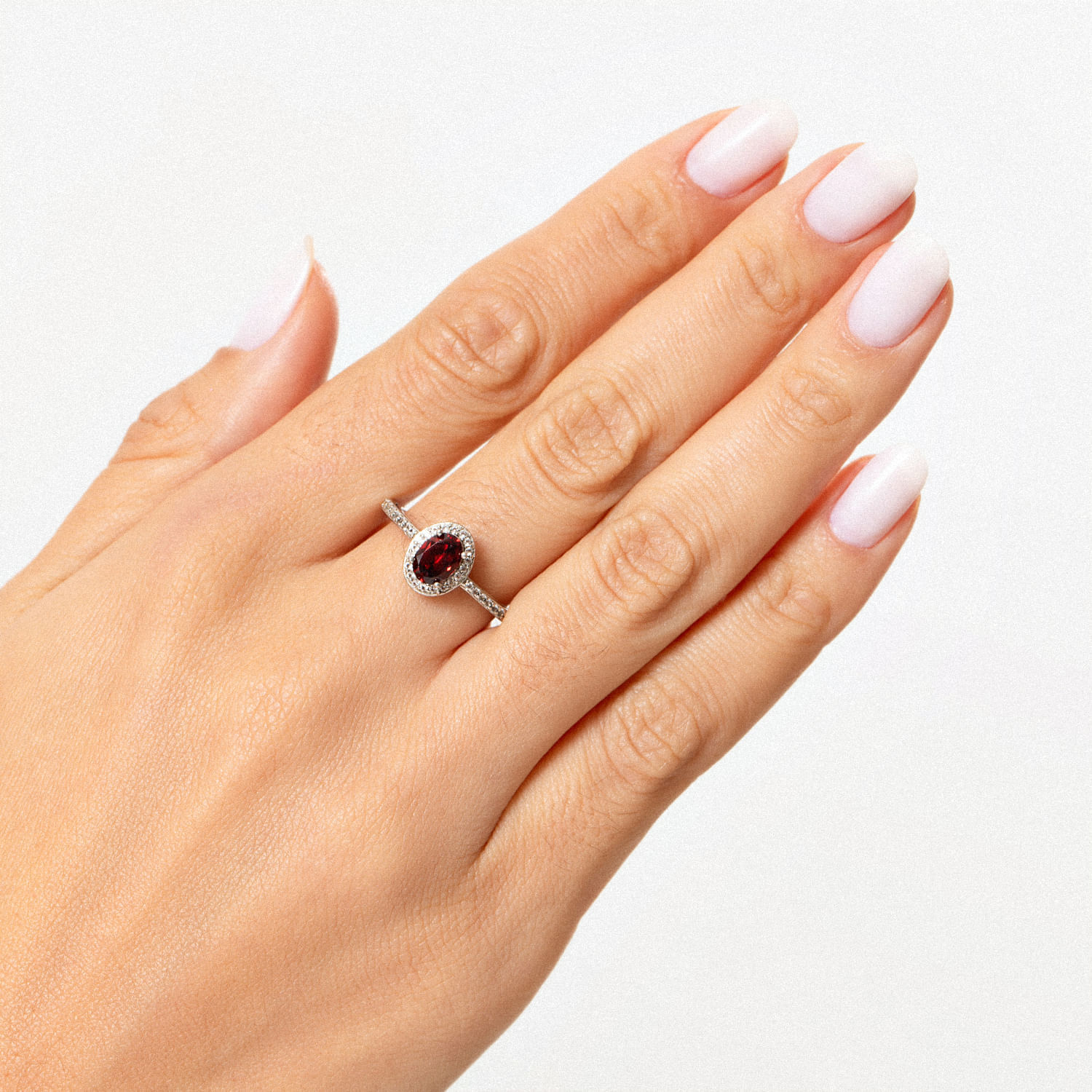 Uma mão elegante com esmalte rosa claro usando um anel de prata com uma grande pedra preciosa vermelha oval cercada por pequenas pedras transparentes.