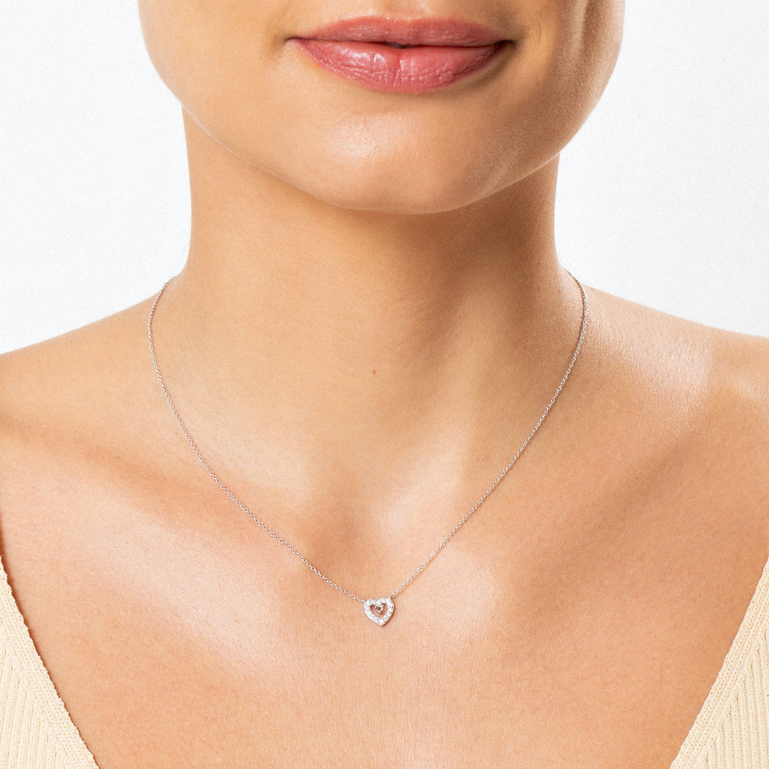 Colar com pingente de coração de prata com pequenas pedras brilhantes, usado por uma pessoa com pele clara e vestido bege.