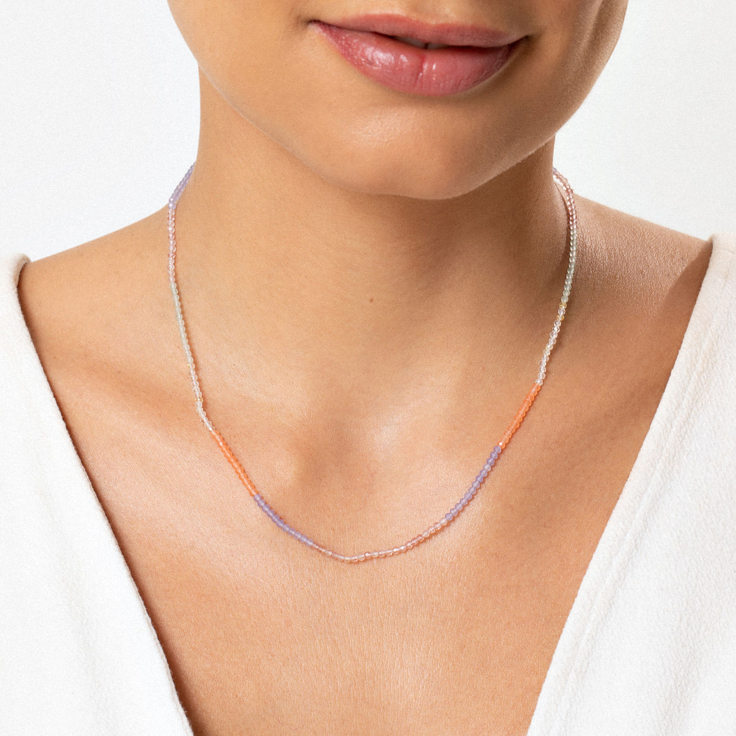 Colar delicado com partes coloridas em tons de roxo, laranja, e prata, usado por uma mulher com pele clara e blusa branca.