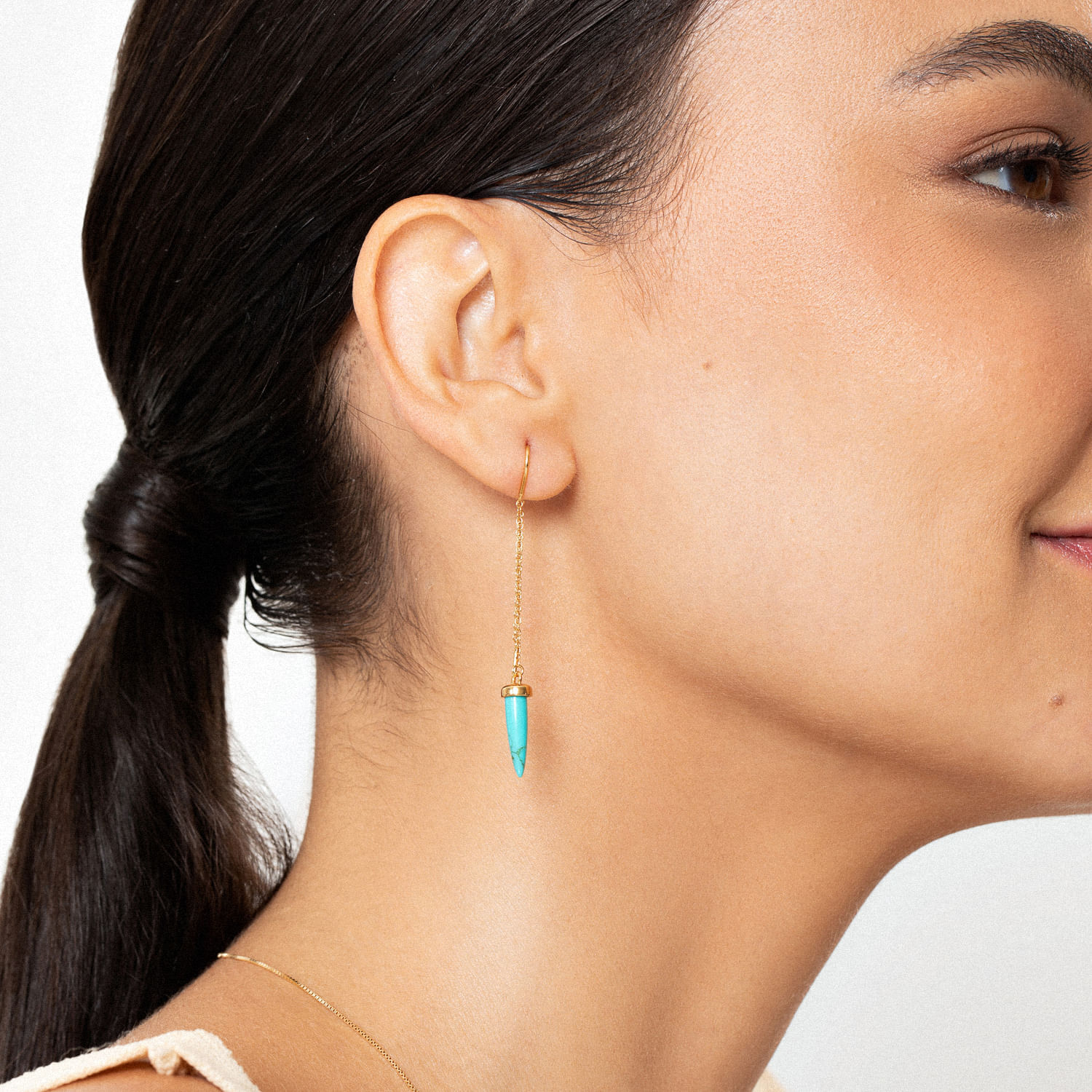 Close de uma mulher usando um brinco de corrente de ouro com um pingente de turquesa, cabelo preso para trás e um fundo neutro.