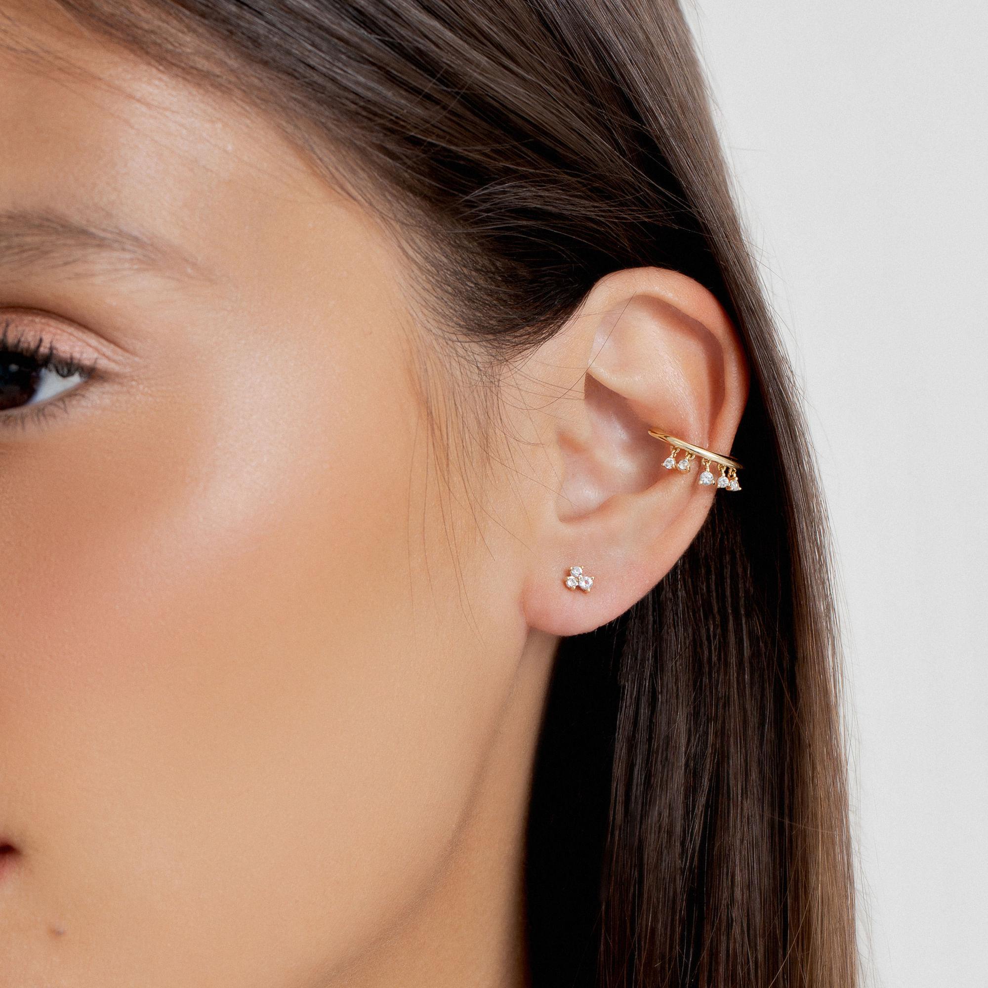 Close da orelha de uma mulher adornada com vários brincos de ouro, incluindo um pequeno pino e um bracelete com cristais pendurados.