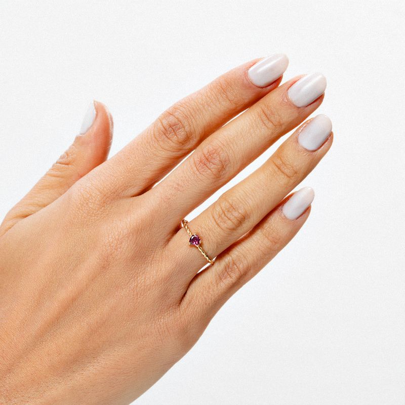 Dedo com unhas brancas e anel delicado com pedra roxa na mão de pele clara contra fundo branco.