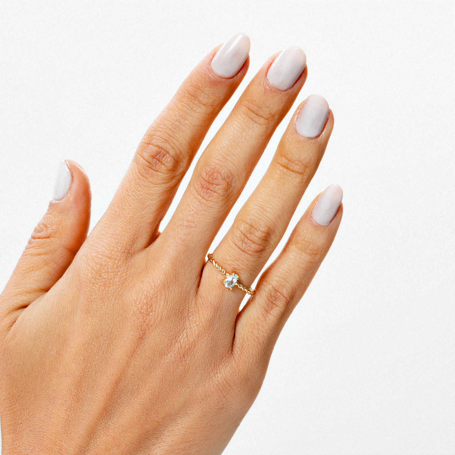 Mão com unhas brancas e um anel dourado com pedra azul claro no dedo indicador.
