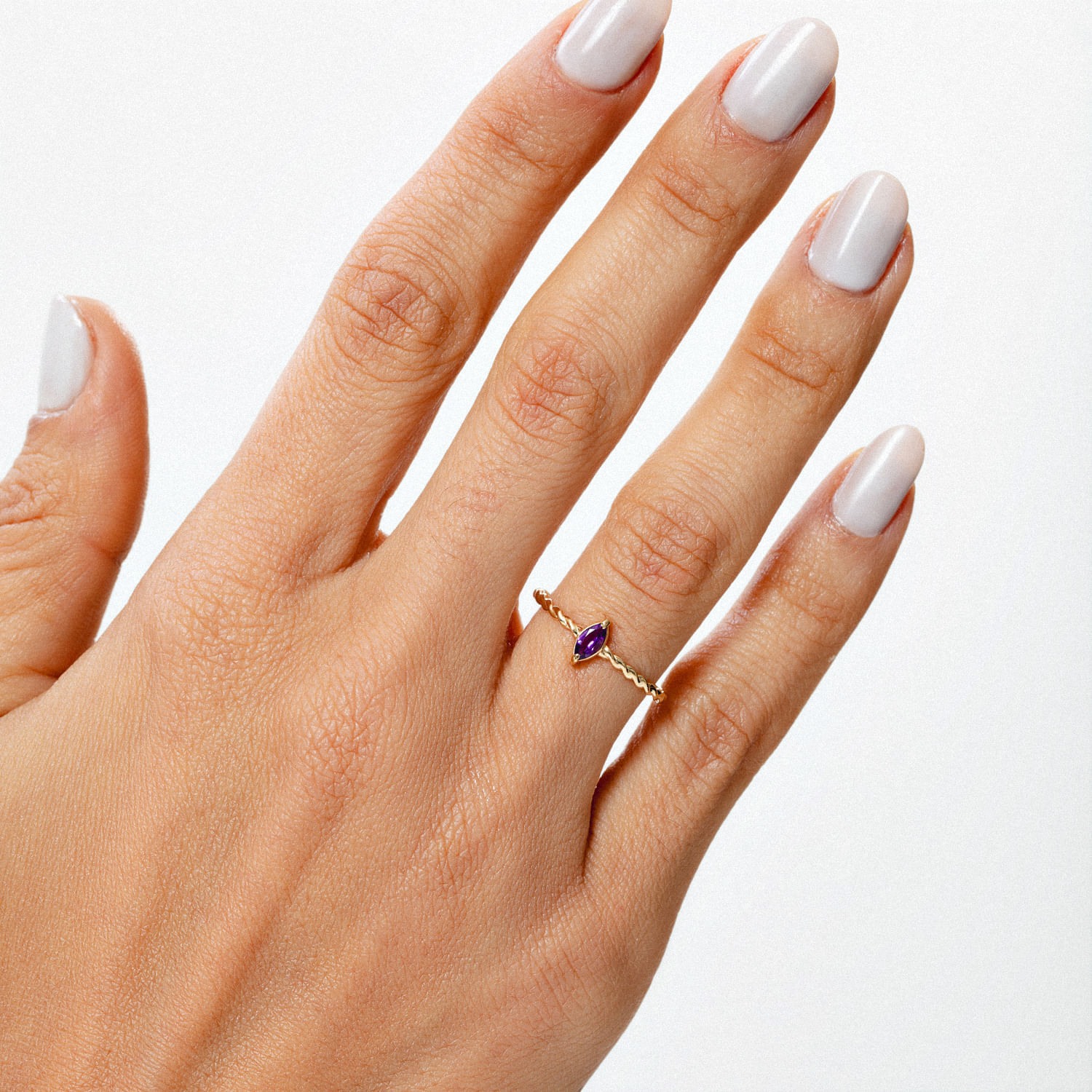 Dedo de uma mão com unhas brancas, usando um anel dourado com pedra roxa na mão contra fundo branco.