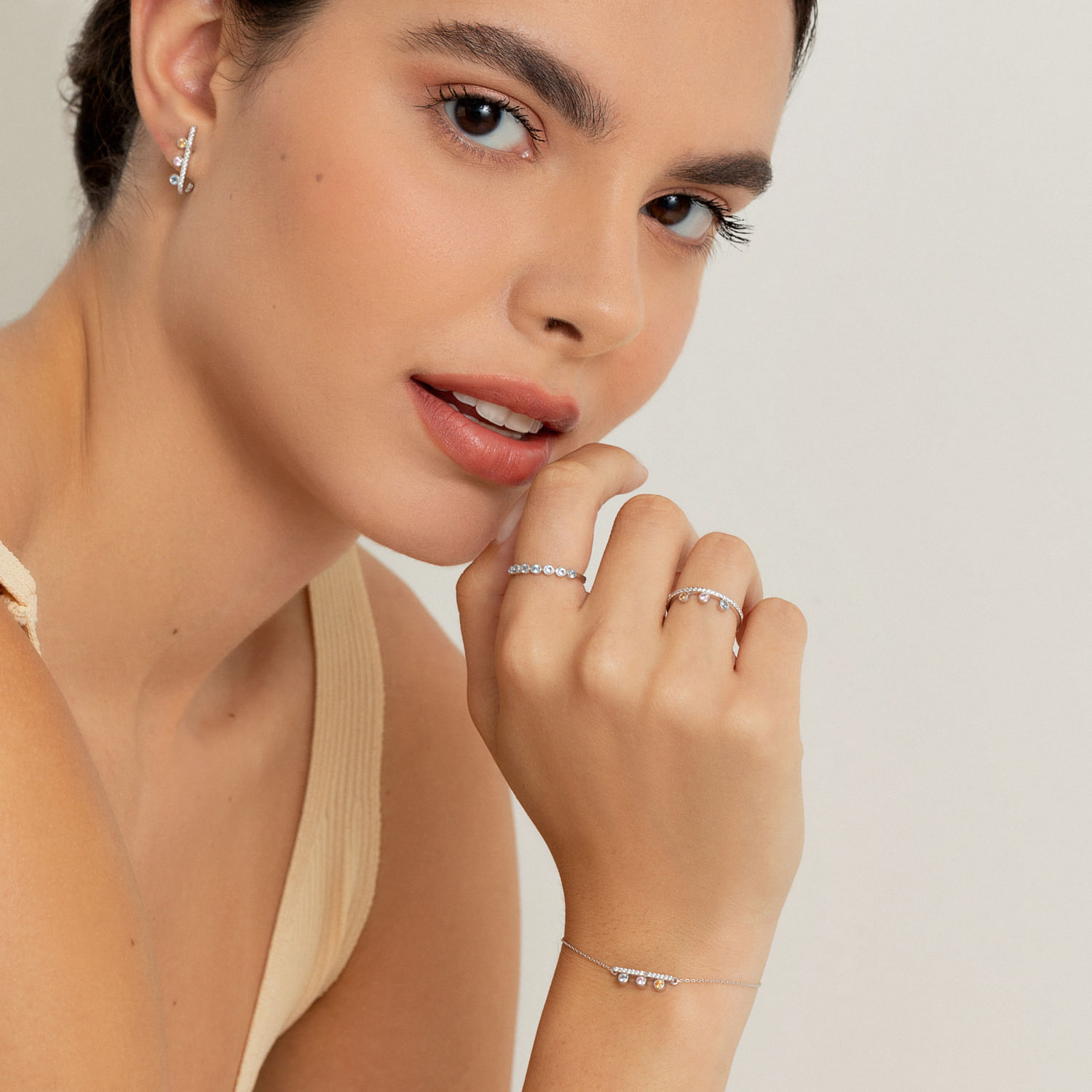 Mulher com brincos, anel, pulseira e anéis com pedras brilhantes, vestindo blusa bege claro, posando com a mão próxima ao rosto.